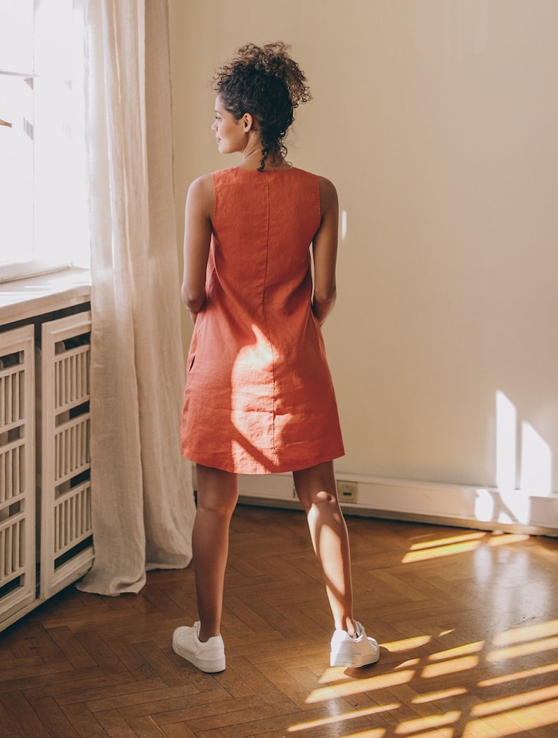 May include: A woman wearing a sleeveless, coral-coloured linen dress and white trainers. She is standing in a room with a window and wooden floors.
