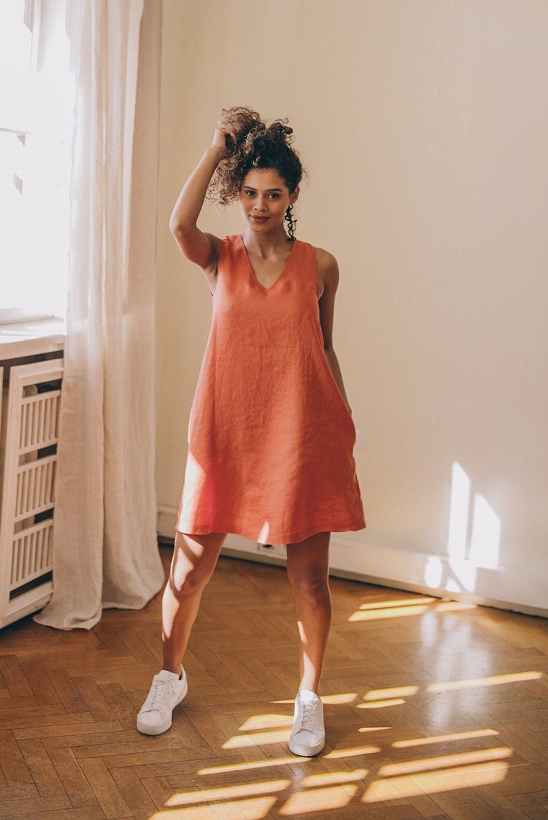May include: A woman wearing a coral linen sleeveless dress with a v-neck and pockets. She is standing on a wooden floor wearing white trainers.
