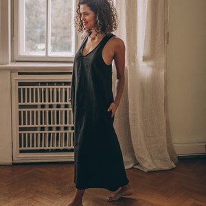 May include: A black, sleeveless maxi dress with a V-neck. The dress has a relaxed fit and pockets. The woman is standing in a room with a window and curtains. The dress is made of a lightweight fabric.