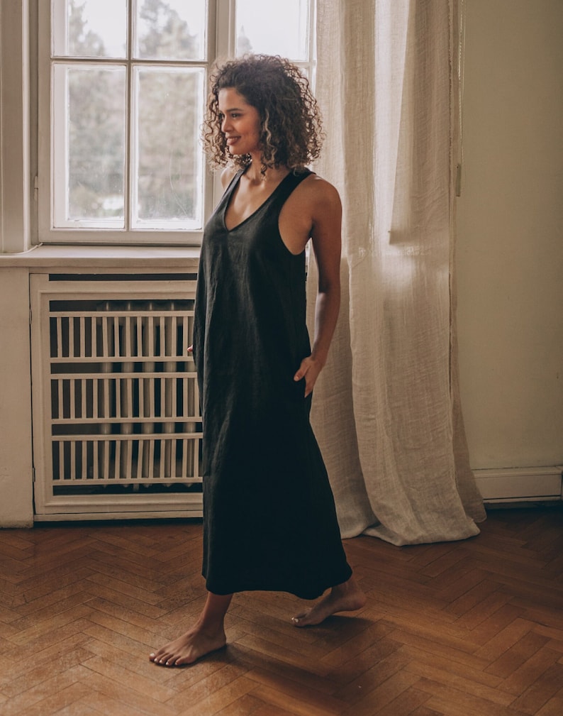 May include: A black, sleeveless maxi dress with a V-neck. The dress has a relaxed fit and pockets. The woman is standing in a room with a window and curtains. The dress is made of a lightweight fabric.