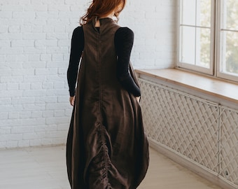 May include: A brown linen dress with a high neckline and a gathered, bubble-style skirt. The dress is worn over a black long-sleeved top. The model is walking away from the camera in a bright room with a white brick wall and a large window.