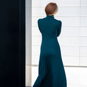 May include: A woman wearing a long, teal-colored dress with a turtleneck. She is standing in front of a white wall with a black door to her left.