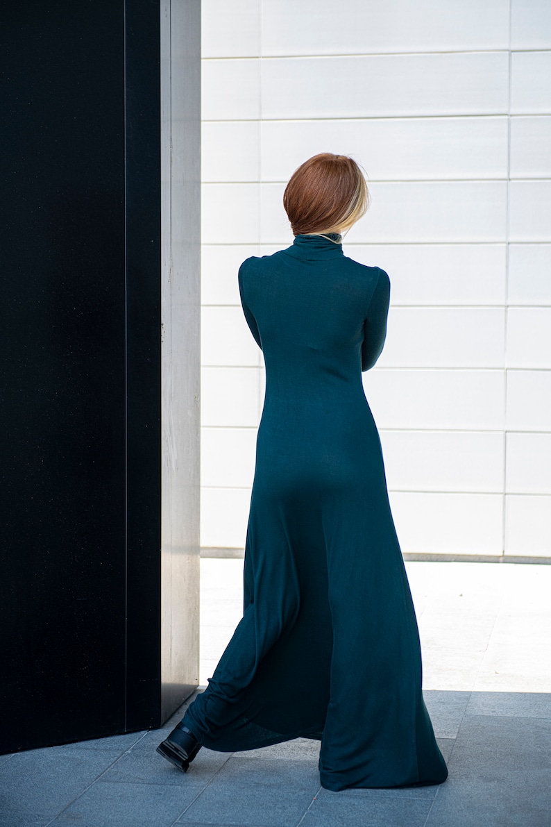 May include: A woman wearing a long, teal-colored dress with a turtleneck. She is standing in front of a white wall with a black door to her left.