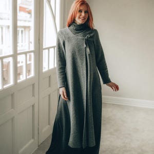 May include: A long-sleeved, charcoal grey wrap-style coat featuring a high turtleneck collar and a side tie closure. The garment is constructed from a textured fabric and extends to a full-length silhouette, layered over a dark, flowing skirt.