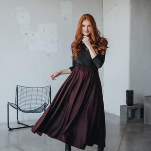 May include: A woman with long red hair wearing a black long-sleeved shirt and a burgundy maxi skirt. She is standing in a room with a white wall and a black metal chair.