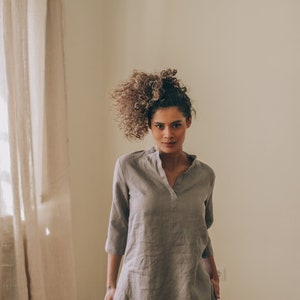 May include: A gray linen tunic dress with a mandarin collar and three-quarter sleeves. The dress has a relaxed fit and two front pockets. The model is wearing white shorts underneath. The dress is approximately 36 inches long.