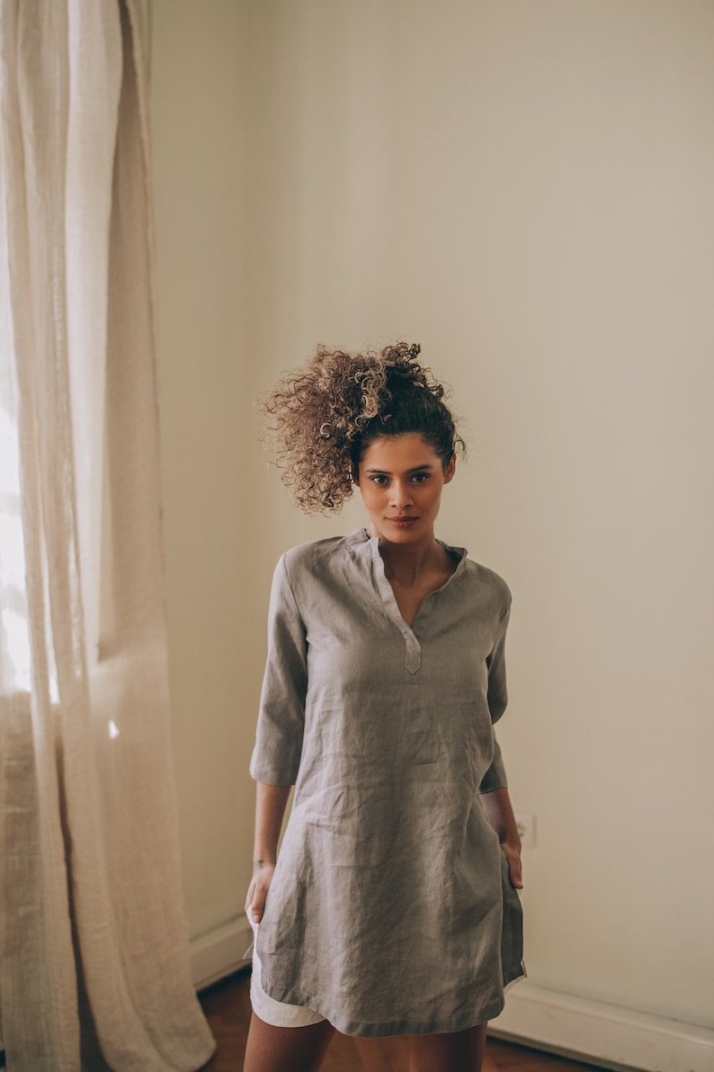 May include: A gray linen tunic dress with a mandarin collar and three-quarter sleeves. The dress has a relaxed fit and two front pockets. The model is wearing white shorts underneath. The dress is approximately 36 inches long.