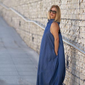 Puede incluir: Una mujer con un vestido de mezclilla azul de cuello alto y corte holgado y fluido. Est&aacute; de pie frente a una pared de piedra y lleva gafas de sol.