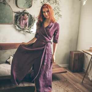 Puede incluir: Un vestido cruzado de color ciruela con cuello en V y cintura anudada. El vestido tiene mangas tres cuartos y una falda fluida. La modelo lleva sandalias marrones. El fondo incluye un banco de madera, espejos y vegetaci&oacute;n colgante.
