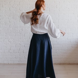 May include: A white linen blouse with long, billowing sleeves and a navy blue linen skirt. The skirt is a wrap style, and the outfit is paired with white sneakers. The background is a white brick wall.