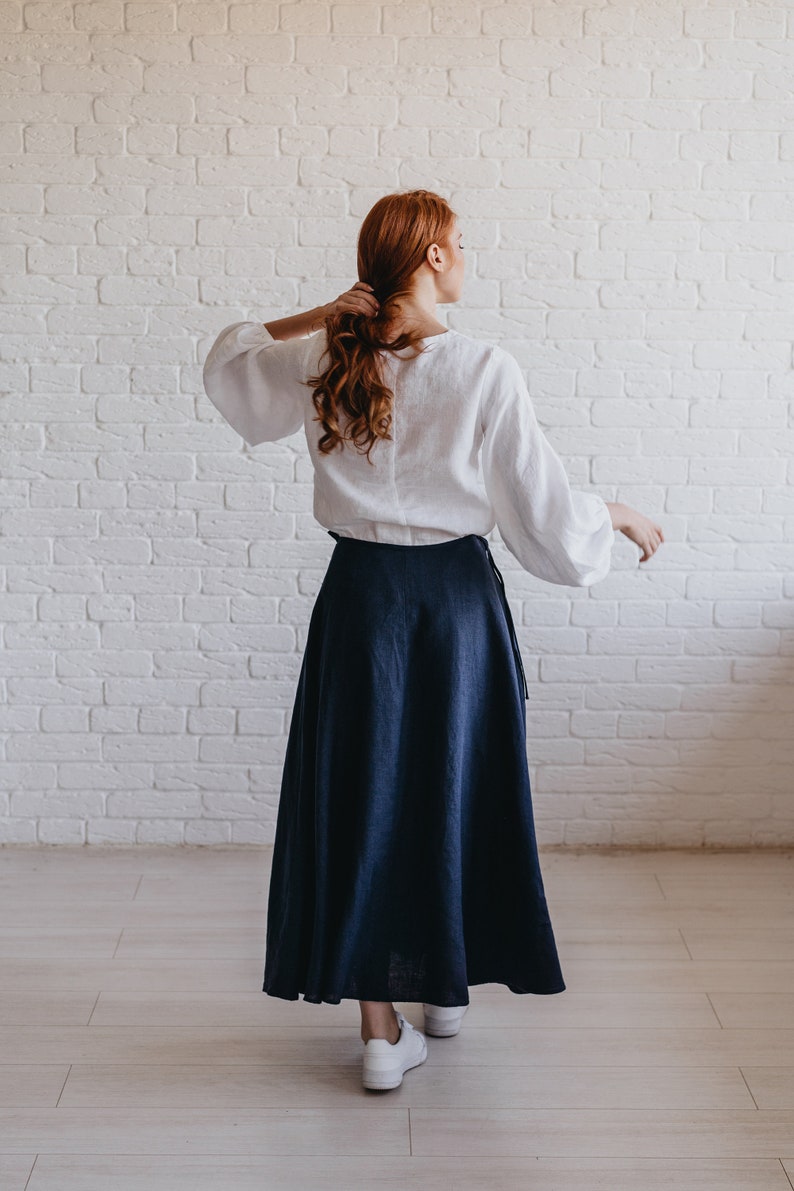 May include: A white linen blouse with long, billowing sleeves and a navy blue linen skirt. The skirt is a wrap style, and the outfit is paired with white sneakers. The background is a white brick wall.