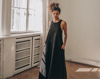 May include: A sleeveless, floor-length black dress with a relaxed fit. The dress features a high neckline and pockets. The model is standing in a room with a large window and hardwood floors. The dress is made of a lightweight fabric.