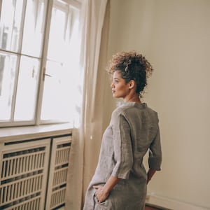 May include: A woman wearing a light gray linen tunic and matching pants. The tunic has a mandarin collar and three-quarter sleeves. The outfit is simple and elegant, suitable for casual or semi-formal wear. The woman is standing near a window with natural light.