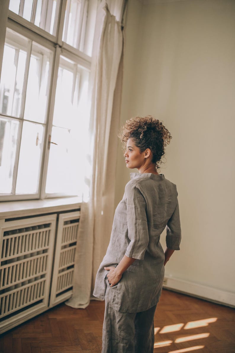 May include: A woman wearing a light gray linen tunic and matching pants. The tunic has a mandarin collar and three-quarter sleeves. The outfit is simple and elegant, suitable for casual or semi-formal wear. The woman is standing near a window with natural light.