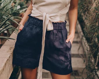 May include: A beige wrap top with a tie closure paired with navy blue shorts. The shorts have an elastic waistband and pockets. The outfit is made of a lightweight fabric, perfect for warm weather. The background features lush greenery.