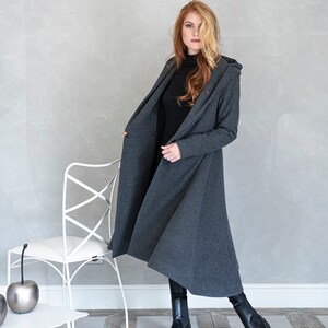 May include: A long, gray hooded cardigan with a slightly flared hem. The cardigan is open and the model is wearing a black turtleneck underneath.