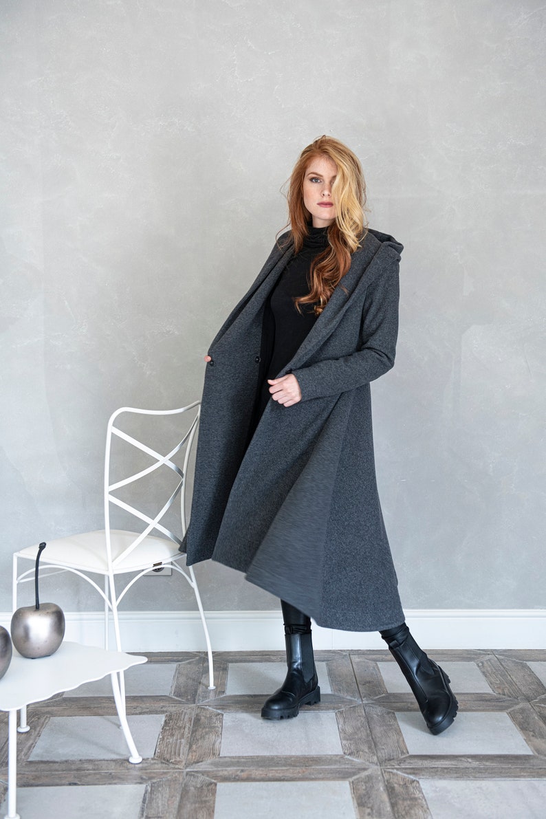 May include: A long, gray hooded cardigan with a slightly flared hem. The cardigan is open and the model is wearing a black turtleneck underneath.