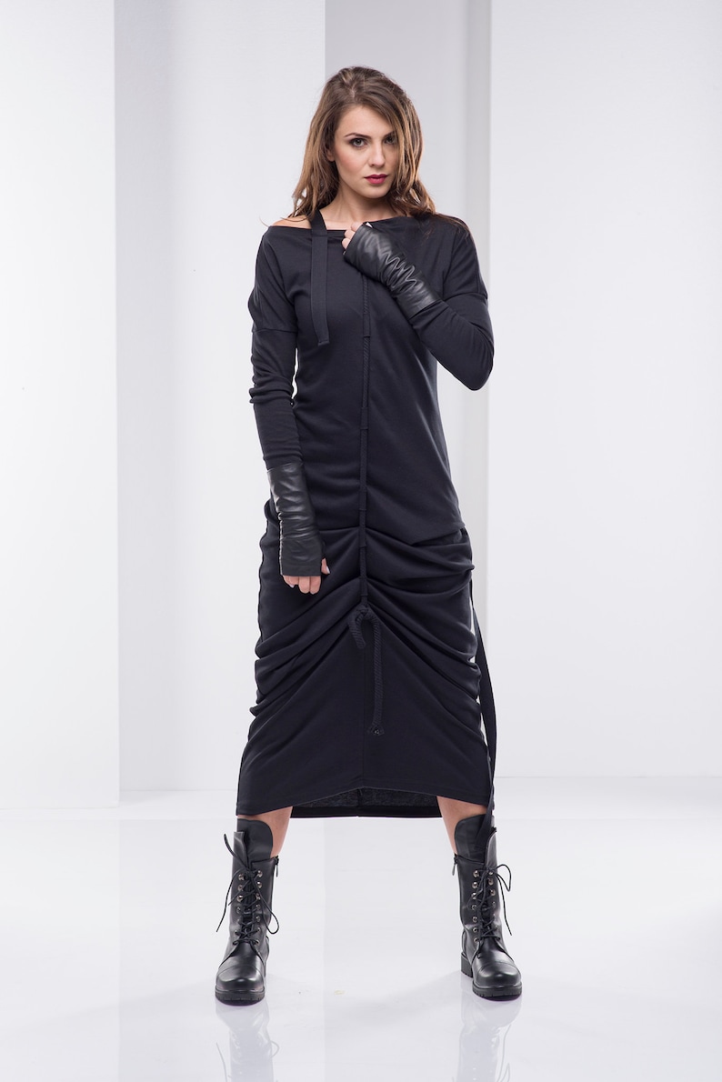 May include: A black long-sleeved dress with a gathered skirt and a long, thin tie at the waist. The dress has a cowl neck and is worn with black leather gloves and black lace-up boots.