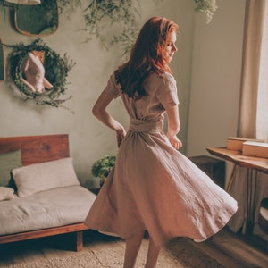 May include: A light pink linen dress with a short-sleeve design and a matching belt. The dress is knee-length and has a flowing skirt. The person is wearing brown sandals. The room has a natural, earthy aesthetic with plants and wooden furniture.