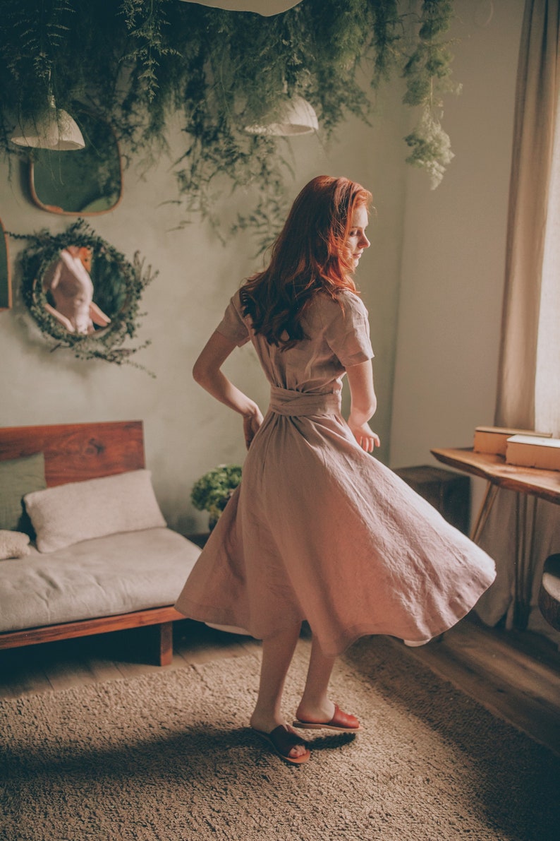 May include: A light pink linen dress with a short-sleeve design and a matching belt. The dress is knee-length and has a flowing skirt. The person is wearing brown sandals. The room has a natural, earthy aesthetic with plants and wooden furniture.