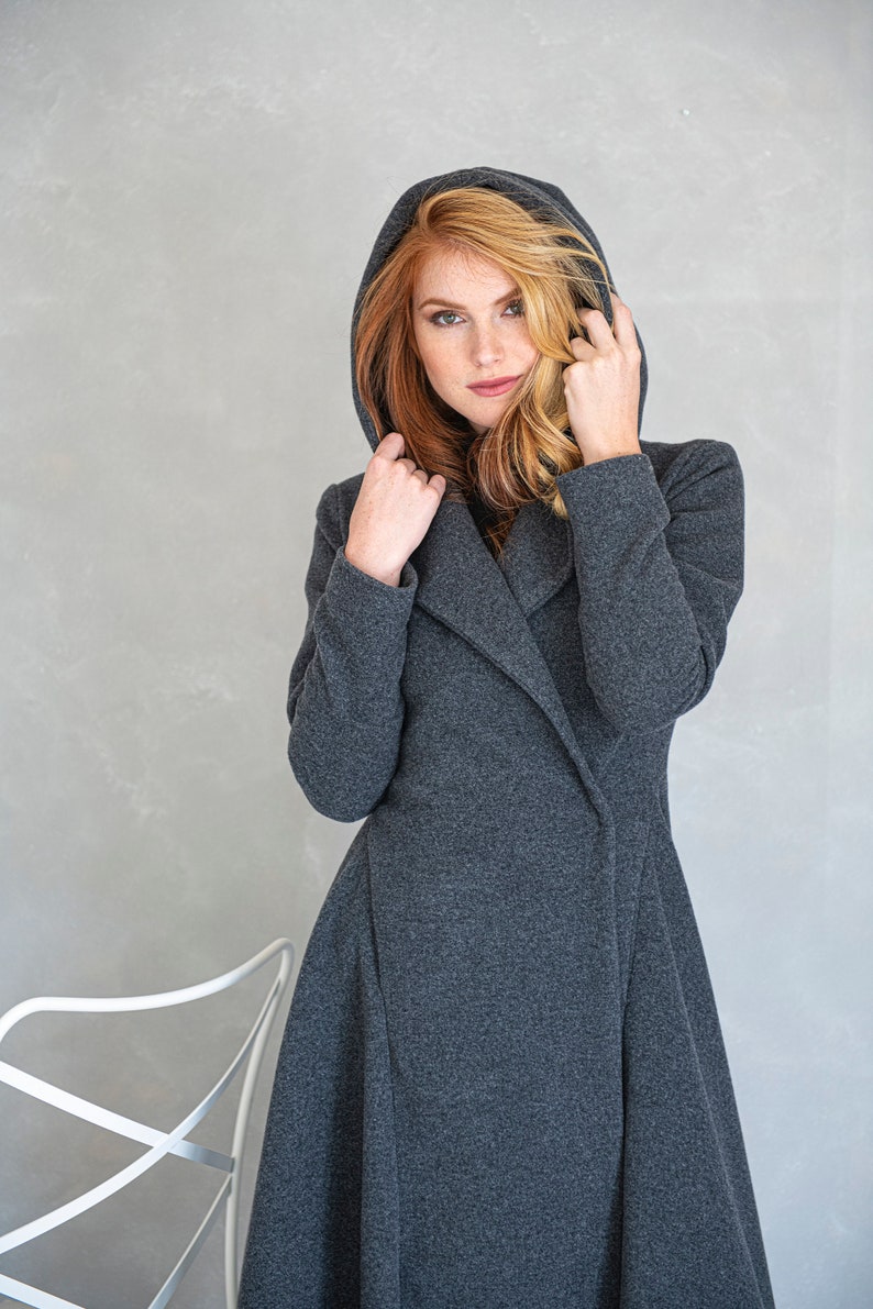 May include: A woman wearing a long, dark gray hooded coat with a shawl collar. The coat has a fitted bodice and a flowing skirt.