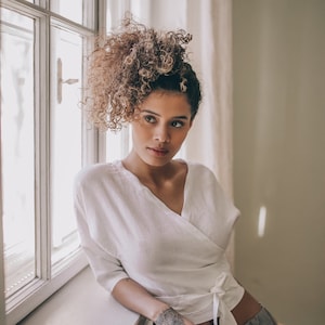May include: A white wrap top with a tie closure. The top is paired with gray pants. The model is leaning against a window frame. The model has curly hair and a tattoo on their arm.