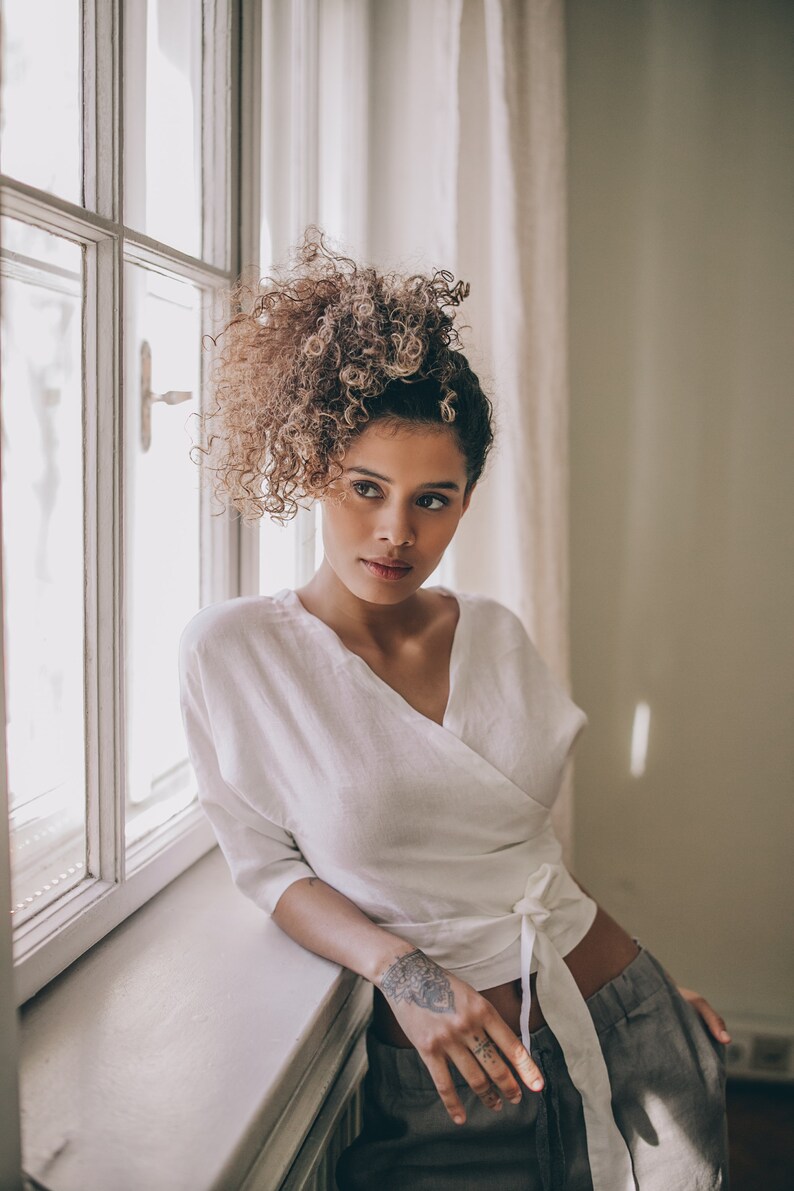 May include: A white wrap top with a tie closure. The top is paired with gray pants. The model is leaning against a window frame. The model has curly hair and a tattoo on their arm.