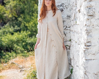 May include: A beige linen outfit consisting of a long-sleeved, button-down duster and a matching skirt. The outfit is worn by a person standing next to a white stone wall, with green foliage in the background. The outfit is made of a lightweight fabric.