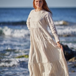 Może przedstawiać: Kobieta z długimi, czerwonymi włosami w białej lnianej sukience stoi na piaszczystej plaży z falami oceanu w tle.