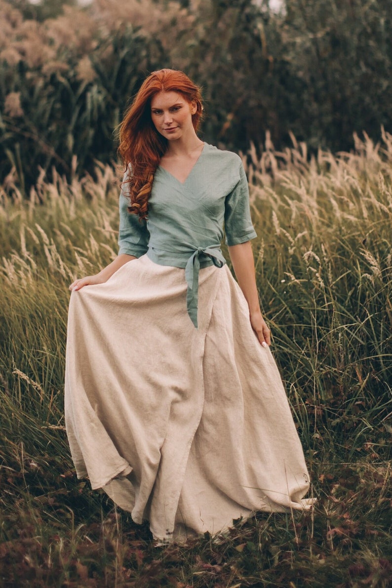 May include: A woman wearing a sage green wrap top with three-quarter sleeves and a beige wrap skirt. The outfit is made of linen and is suitable for warm weather. The model is standing in a field of tall grass.
