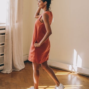 May include: A woman wearing a sleeveless, coral-coloured linen dress and white trainers. She is standing in a room with hardwood floors and a white wall.