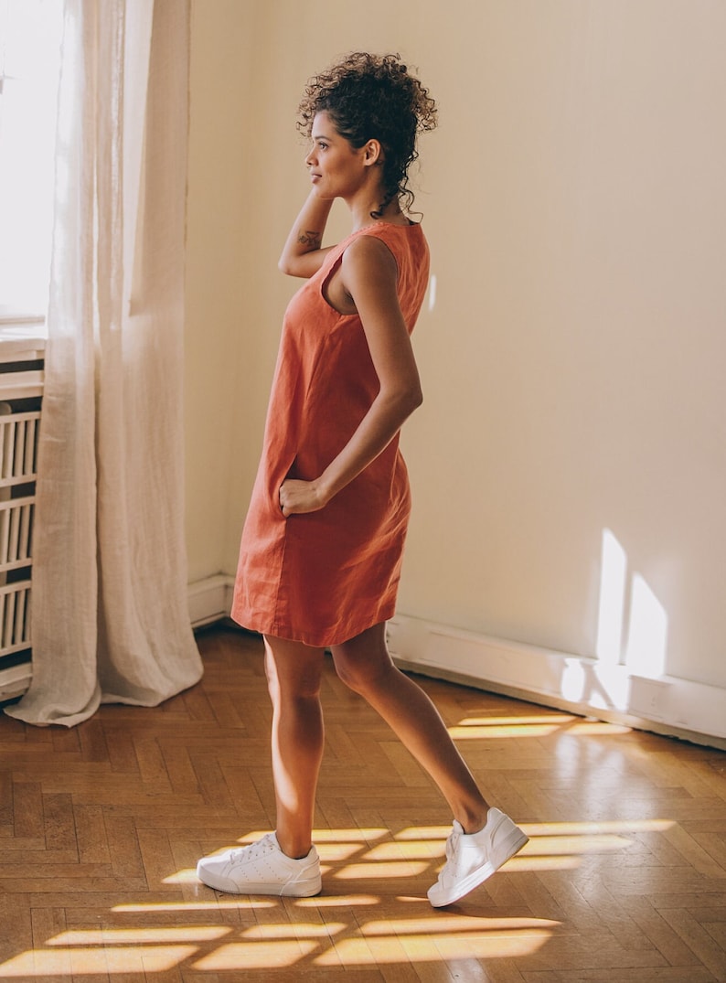 May include: A woman wearing a sleeveless, coral-coloured linen dress and white trainers. She is standing in a room with hardwood floors and a white wall.