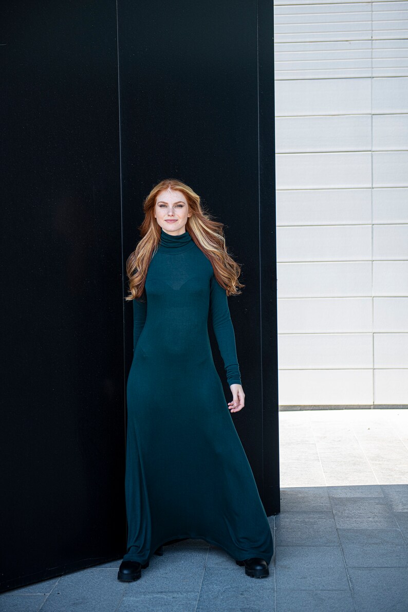 May include: A woman with long, red hair is wearing a long, teal green dress. She is standing in front of a black and white wall.