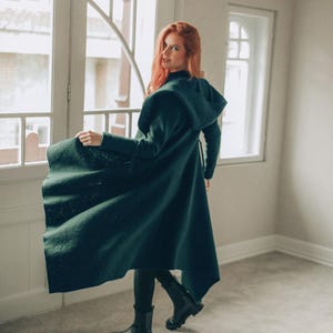 May include: A long, dark green coat with a high collar and hood. The coat is worn with black boots and leggings. The person is standing near a window with white trim. The coat appears to be made of a thick, textured fabric.