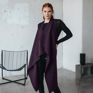 Könnte beinhalten: Eine lange, drapierte Weste in Pflaumenfarbe. Die Weste wird über einem schwarzen Langarm-Top und einer schwarzen Hose getragen. Das Model steht in einem modernen, minimalistischen Raum mit einem schwarzen Stuhl und einer dekorativen schwarzen Vase.