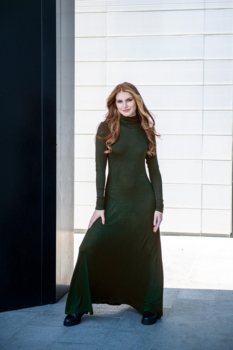 May include: A woman wearing a long, dark green, turtleneck dress with long sleeves. She is standing in front of a white wall and a black wall.