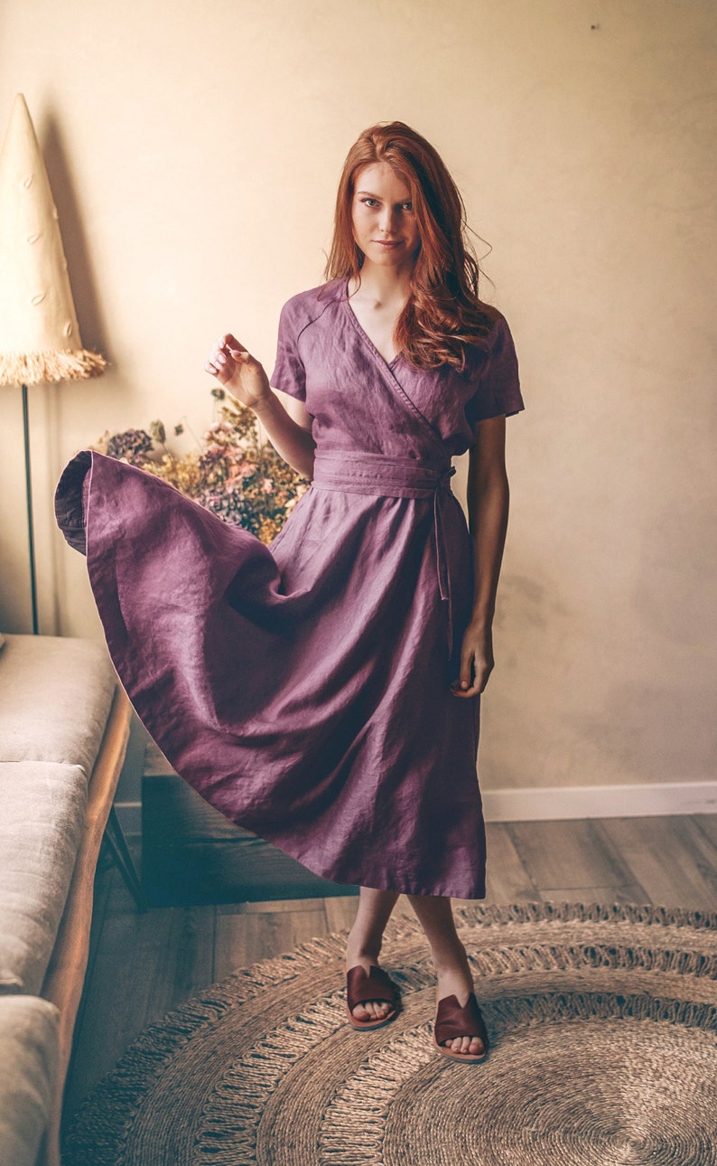 May include: A mauve wrap dress with short sleeves and a matching belt. The dress is made of a flowing fabric and is paired with brown sandals. The model is standing on a round jute rug.