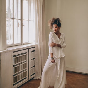 May include: A woman's white linen outfit, consisting of a long-sleeved top and wide-leg pants, is shown in a well-lit room. The outfit has a relaxed fit and is paired with white sneakers. The room features a large window and wooden floors.