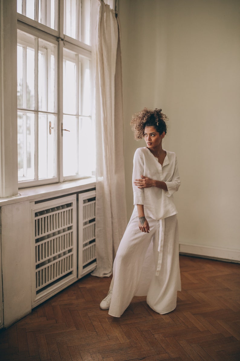 May include: A woman's white linen outfit, consisting of a long-sleeved top and wide-leg pants, is shown in a well-lit room. The outfit has a relaxed fit and is paired with white sneakers. The room features a large window and wooden floors.