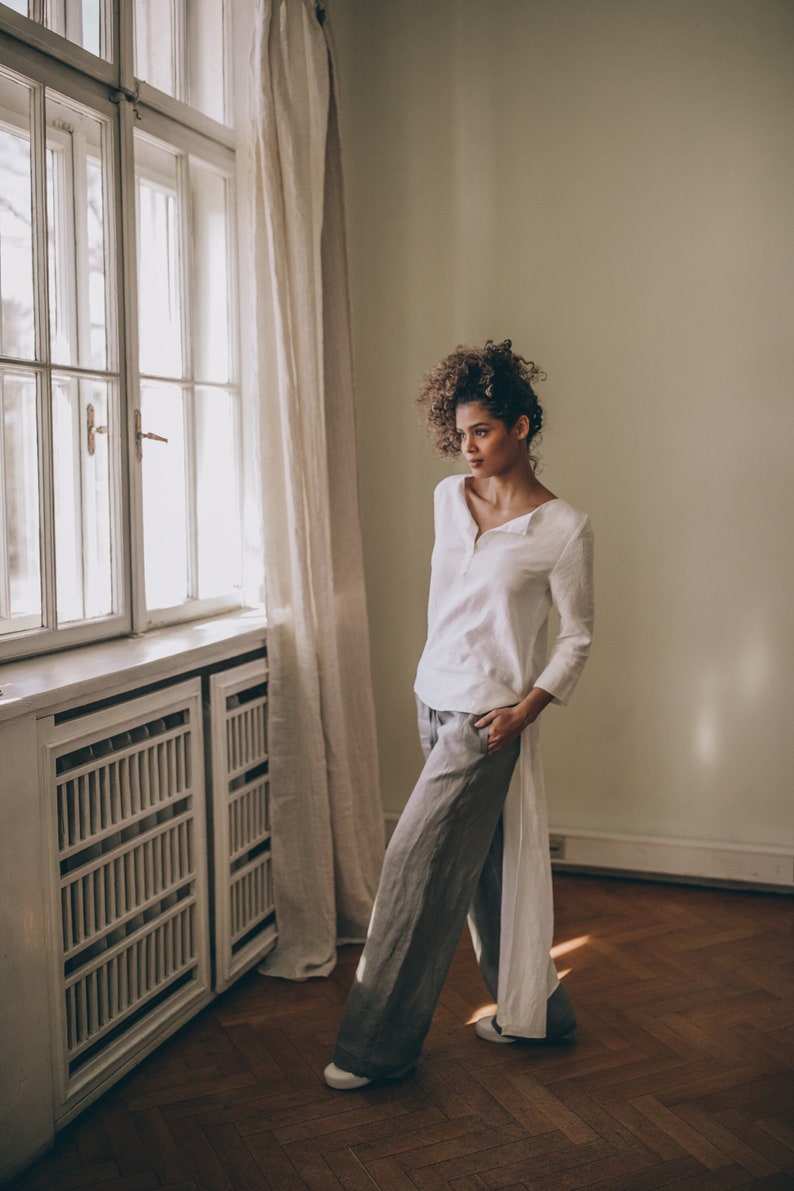 May include: A woman wearing a white linen top with a long, split back and gray linen pants. The outfit is paired with white shoes. The setting is a room with a large window, curtains, and a wooden floor.
