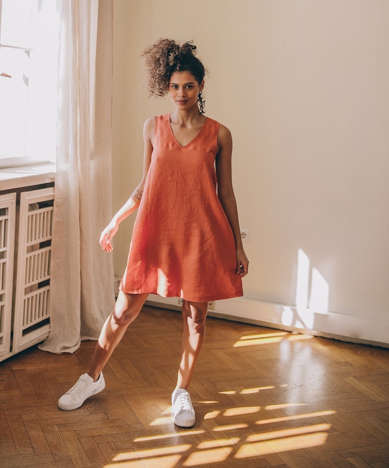 May include: A woman wearing a coral sleeveless linen dress and white trainers stands in a room with hardwood floors.