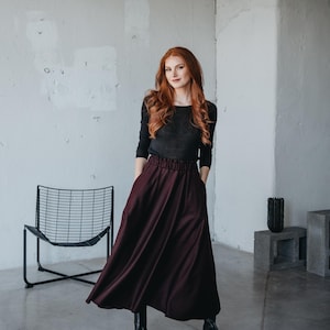 May include: A woman wearing a black long-sleeved shirt and a burgundy maxi skirt. She is standing in a room with a white wall and a black metal chair.