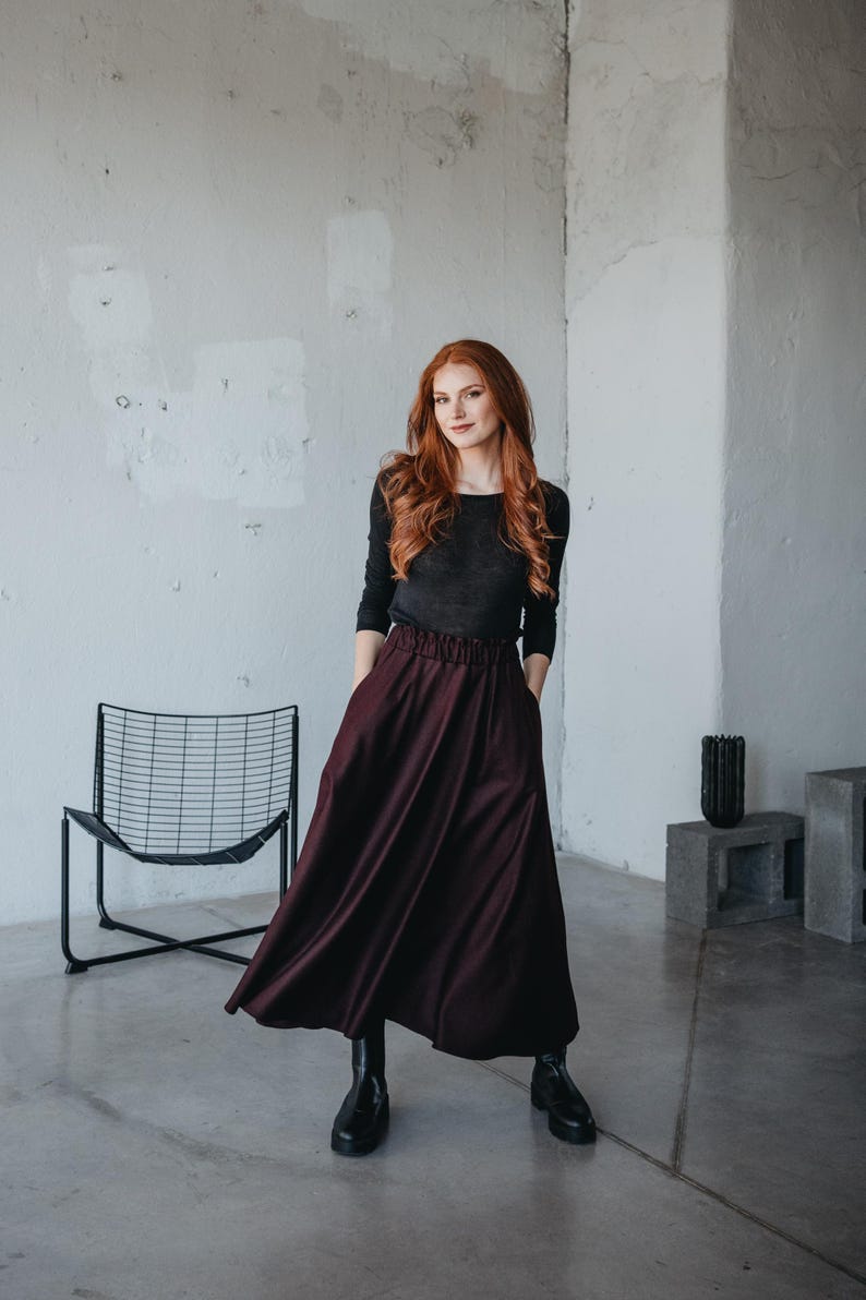 May include: A woman wearing a black long-sleeved shirt and a burgundy maxi skirt. She is standing in a room with a white wall and a black metal chair.