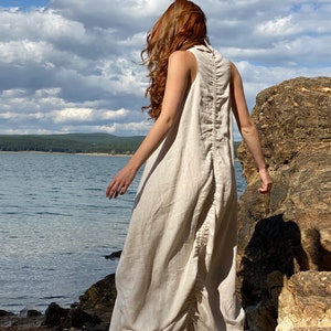 May include: A sleeveless, floor-length beige linen dress. The dress features a high collar and a gathered, ruched design down the back. The model is standing near a lake with a rocky shoreline and a cloudy sky in the background.