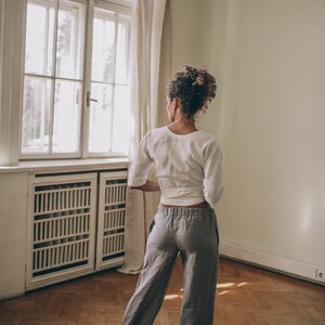 May include: A person wearing a white wrap top and gray linen pants. The outfit is paired with white sneakers. The person is standing in a room with a large window and hardwood floors. The room has a neutral color palette.