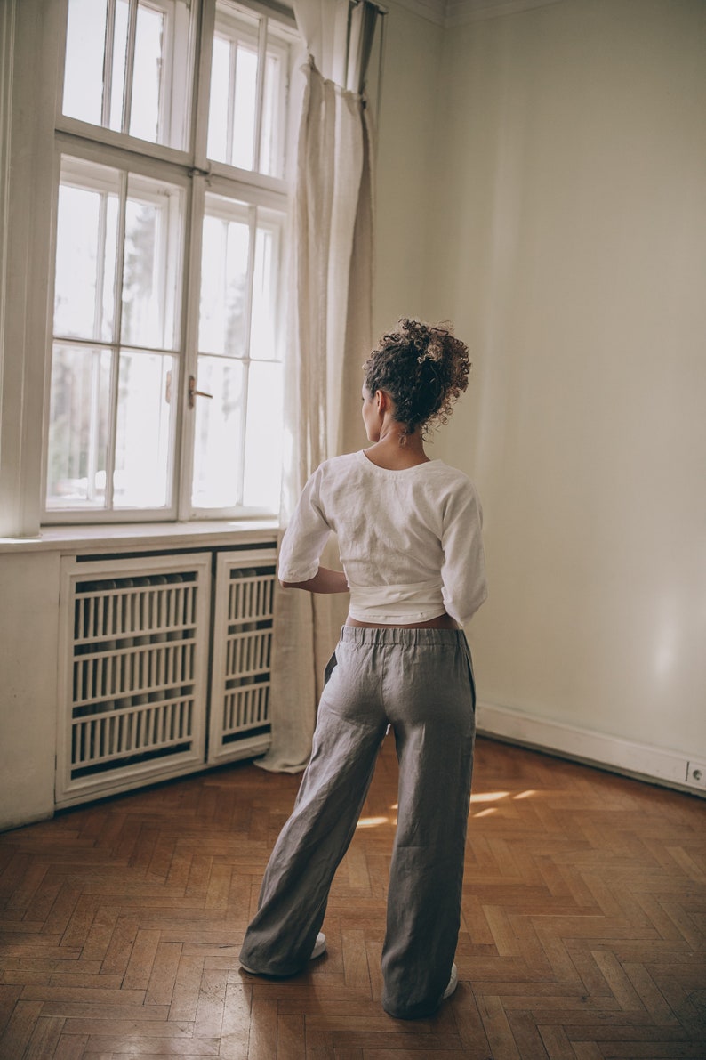 May include: A person wearing a white wrap top and gray linen pants. The outfit is paired with white sneakers. The person is standing in a room with a large window and hardwood floors. The room has a neutral color palette.