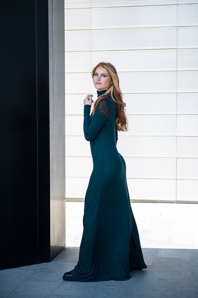 May include: A woman wearing a long, dark green maxi dress with a high neck and long sleeves. She is standing in front of a white wall and a black wall.
