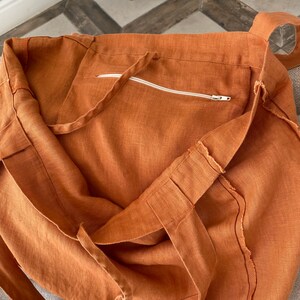 May include: A large, rust-colored linen tote bag with a zippered pocket on the inside. The bag has a raw edge finish and is made of natural linen.