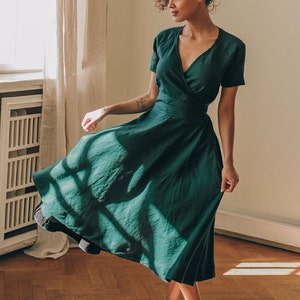 May include: A dark green wrap dress with short sleeves and a midi-length skirt. The dress features a V-neckline and a fitted waist. The person in the image is wearing white sneakers and standing on a wooden floor.