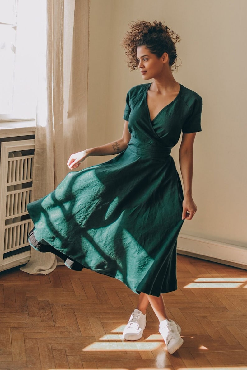 May include: A dark green wrap dress with short sleeves and a midi-length skirt. The dress features a V-neckline and a fitted waist. The person in the image is wearing white sneakers and standing on a wooden floor.